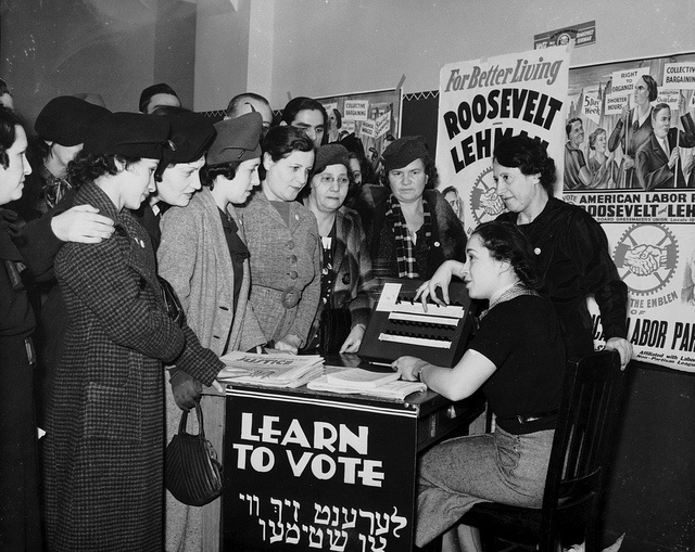Women surrounded by posters in English and Yiddish supporting Franklin D. Roosevelt, Herbert H. Lehman, and the American Labor Party teach other women how to vote, 1935